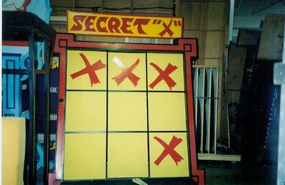 A game display titled 'SECRET X' featuring a yellow board with red crosses and an empty center square, set in a storage area.