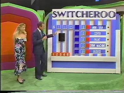 A host and a model stand next to a game board for the game 'Switcheroo' on a game show, with colorful graphics and various items displayed.