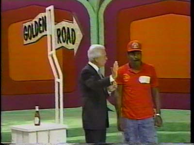 A male contestant in a red shirt stands next to the host of The Price is Right, with a 'Golden Road' sign in the background.