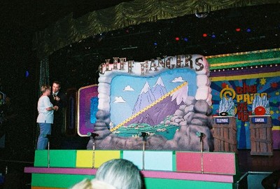 A colorful game show set featuring a mountain-themed backdrop and a stage with two podiums labeled 'Contestant' and 'Host'.
