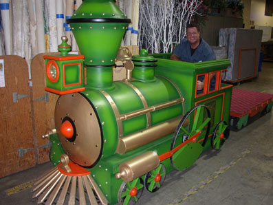 A colorful toy train prop with a smiling man sitting inside it, set against a backdrop of studio materials.