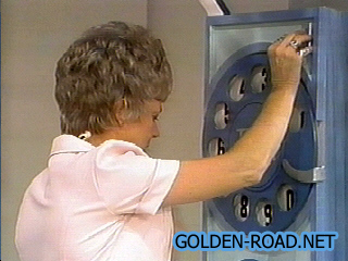 A woman operating a game board on a game show, with a circular dial and numbers displayed.