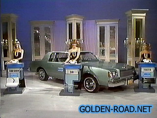 A staged scene from 'The Telephone Game' featuring a green car and three podiums with models holding telephones in front of them.