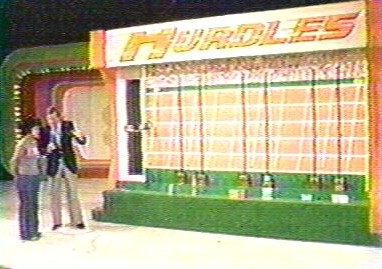 A television game show set featuring a large board labeled 'Hurdles' with a host and a contestant standing in front.