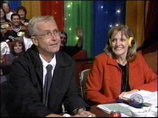 Two individuals sitting at a desk with microphones and headsets during a game show, with a colorful backdrop and audience visible in the background.