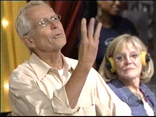 A man, wearing glasses and a light-colored shirt, is gesturing with three fingers raised while speaking, with a woman wearing headphones seated beside him, looking on.
