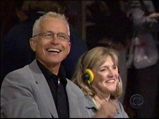 Two smiling individuals, one wearing glasses and a light gray blazer, and Kathy Greco wearing headphones, enjoying a moment during a live game show taping.