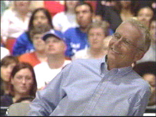 A smiling male participant in a light blue shirt sitting in front of a live audience, with both men and women visible in the background.