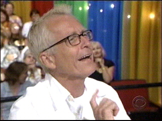 A smiling man wearing glasses reacts excitedly while watching The Price is Right, with a colorful background of curtain panels and audience members visible.