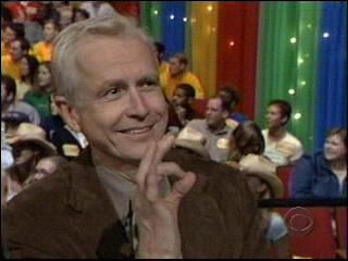 A smiling man in a brown jacket gestures with his fingers while seated in a colorful audience backdrop with various people visible around him.