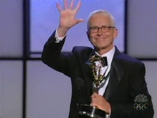 A person in a tuxedo smiles and waves while holding an Emmy award on stage.