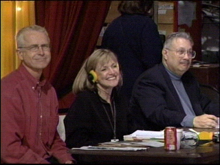 Three individuals sitting at a table during a production, one wearing headphones and smiling, another with glasses, and a third with gray hair, all appearing engaged.