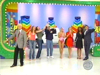 A group of people on stage celebrating during a taping of The Price is Right, with colorful background decorations.