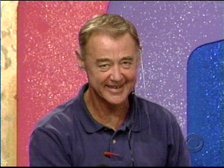 Bob Chic in a blue polo shirt stands in front of a colorful, sparkly background. He appears cheerful and engaged, with a microphone attached to his collar.