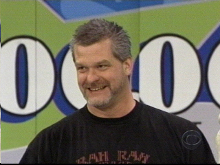 Big Dave with gray hair wearing a black shirt, standing in front of a colorful backdrop featuring the show's logo and numbers.