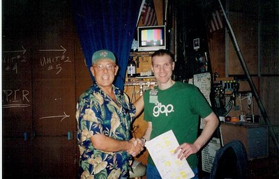 Doug Quick and Doug Bradshaw shaking hands in a backstage area, one wearing a floral shirt and a cap, and the other in a green shirt. Background shows various equipment and a blue curtain.