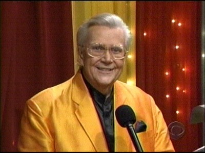 Rod Roddy wearing an orange jacket and glasses stands in front of a colorful backdrop with lights, holding a microphone.