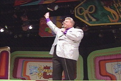 Rod Roddy in a shiny striped suit gestures enthusiastically while standing on stage at The Price is Right, with colorful game show set elements in the background.