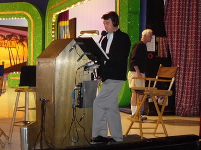 Rich Fields wearing a tuxedo and headphones stands behind a podium at The Price is Right set, with a backdrop displaying colorful decorations.