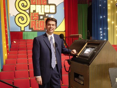 Rich Fields stands next to a podium on set of The Price Is Right, with colorful curtains and a show logo in the background.