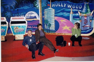 Richard Domabyl, Sue Mcyntire, and Bente Christensen sitting on the floor of a colorful studio set, with game show podiums in the background decorated with a Hollywood theme.