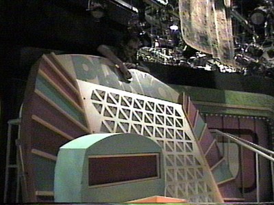A behind-the-scenes view of a game show set, featuring colorful stage props and equipment, with Randy Amasia dropping a chip down the Plinko board.