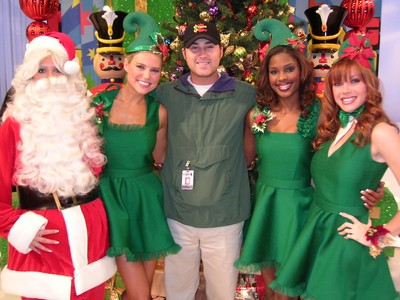 A festive group photo featuring a person dressed as Santa Claus and four individuals, including Scott Schalk, in green holiday outfits, smiling in front of a decorated Christmas tree.