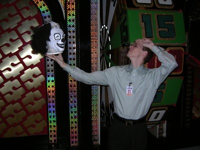 Scott Robinson, a stage crew member poses with a clown wig in front of a colorful game show set, showcasing elements of The Price is Right.