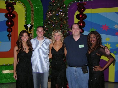 Brandi Sherwood, John Sly, Rachel Reynolds, Marc Green, and Lanisha Cole standing in front of a colorful backdrop decorated for the holidays, featuring a Christmas tree and festive decorations. Two women are dressed in black evening gowns, while the two men are casually dressed.