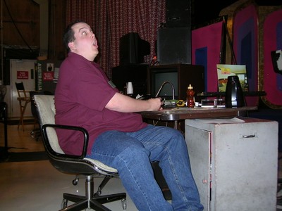 Marc Green sitting at a table with an expressive facial expression, surrounded by various items and equipment, in a studio setting.