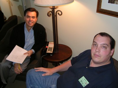 Marc Green and Rich Fields sitting in a room, one holding papers and the other wearing a name tag, with a lamp and a telephone in the background.