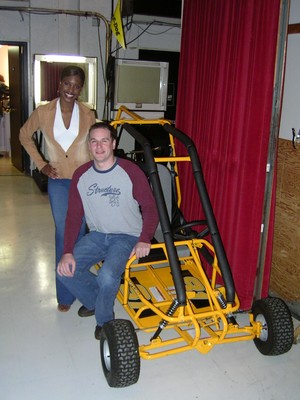 A man wearing a gray sweatshirt and blue jeans sits on a yellow go-kart while a woman in a tan jacket stands next to him, both smiling in a backstage setting.