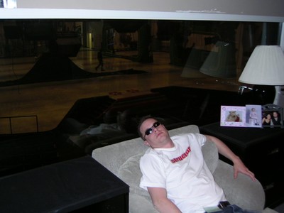 A person lounging on a couch, wearing sunglasses and a casual shirt, with a blurred studio background visible through a window.