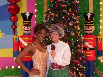 Two women posing in front of a Christmas tree, with colorful nutcracker decorations in the background.