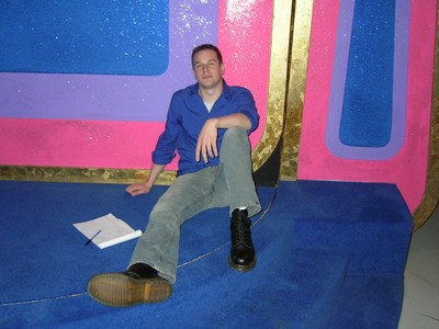 John Sly sitting on blue carpet in front of colorful set designs, wearing a blue shirt and grey pants.