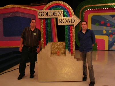 Two individuals posing next to the 'Golden Road' game set on a colorful stage, with glittering decorations in the background.