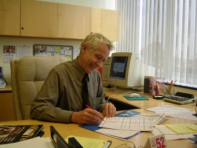 A smiling man in a button-up shirt and tie sits at a cluttered desk, writing on a notepad. In the background, a computer and various office supplies are visible, alongside a window with sheer curtains allowing light to enter.