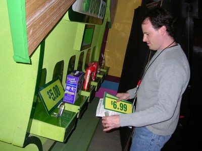 A crew member, Adam Sandler, examines pricing signs on a colorful vending-style setup during a production of The Price is Right.