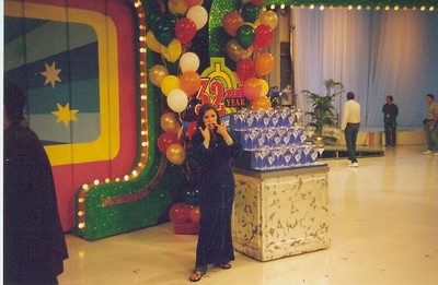 A woman posing with a playful expression in front of colorful balloons and a display featuring a sign for a 40th anniversary celebration, set in a game show studio.