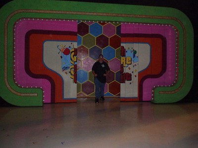 A person standing on a brightly colored television set stage with a hexagonal patterned background resembling a game show set.