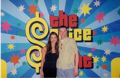 Doug and Brandi Sherwood posing together in front of a colorful backdrop featuring the logo of The Price is Right, with vibrant stars and patterns.