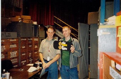 Adam Sandler and Doug Bradshaw standing in a backstage area of a studio, holding a sign with a dollar symbol, surrounded by boxes and storage units.