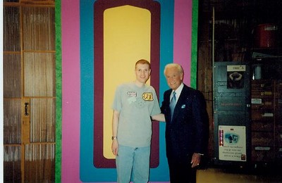 Doug Bradshaw in a shirt and shorts stands next to a well-dressed Bob Barker in a suit, smiling in front of a colorful backdrop.