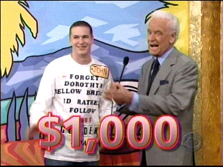 Contestant John smiles while holding a sign on The Price is Right, standing next to host Bob Barker, with a $1,000 prize displayed in front of them.