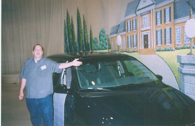 A man standing next to a black car, gesturing towards it, with a mural of a house in the background.