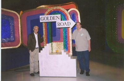 Two men standing next to the Golden Road set piece from The Price is Right, with a colorful background.