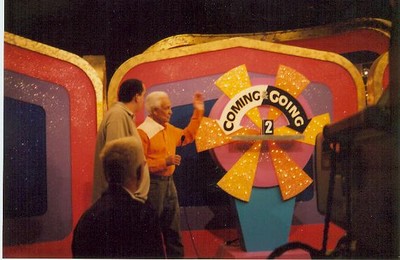 A scene from The Price is Right featuring two men interacting next to a colorful game board labeled 'COMING GOING' with a large spinner.
