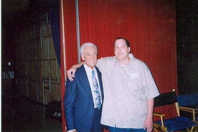 Marc Green standing next to a smiling Bob Barker in a suit, against a red backdrop, inside a studio setting.