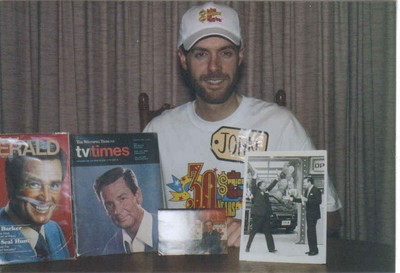 A man wearing a cap and a t-shirt poses at a table with magazines and a picture related to a game show.