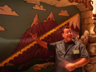 Randy West gestures enthusiastically in front of a colorful mural depicting mountains and clouds.